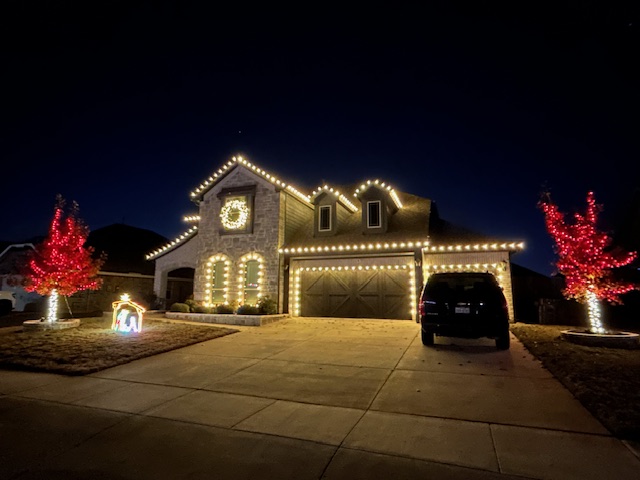 House with dormers, garage doors, and roofline lined with warm white Christmas lights. Trees with red string lights in Forney, TX